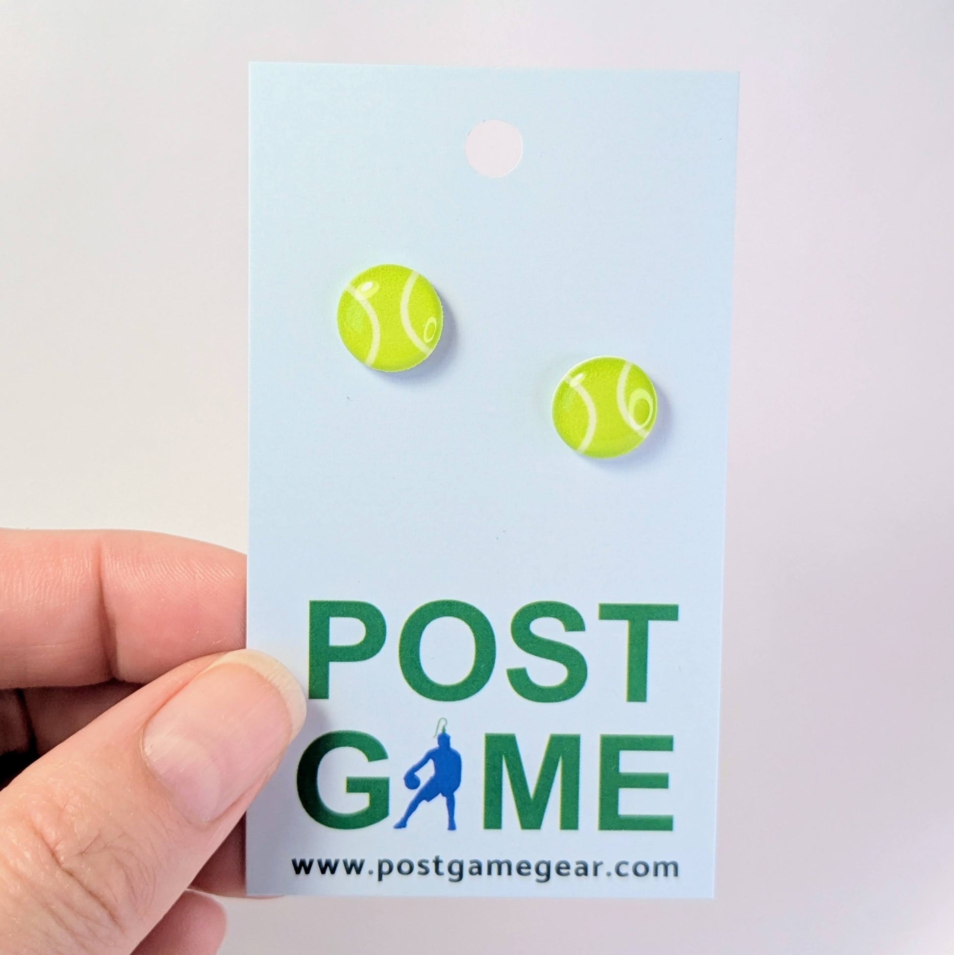 Tennis ball earrings on a 'POST GAME' branded card held by a hand.
