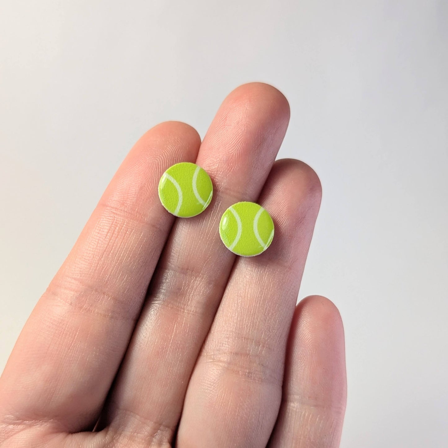 Tennis ball-shaped earrings held between fingers against a plain background