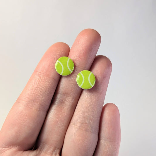 Tennis ball-shaped earrings held between fingers against a plain background