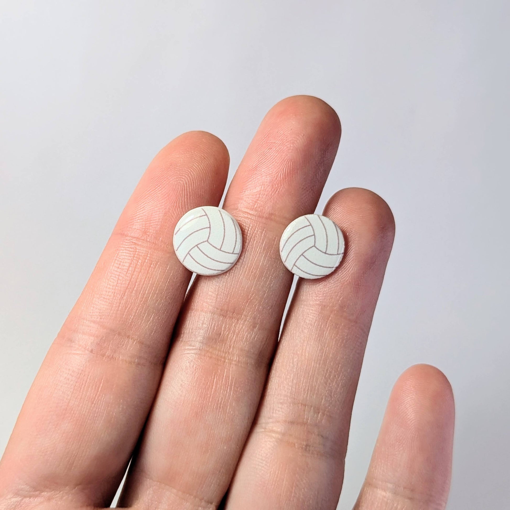 White volleyball-shaped earrings held between fingers against a light gray background