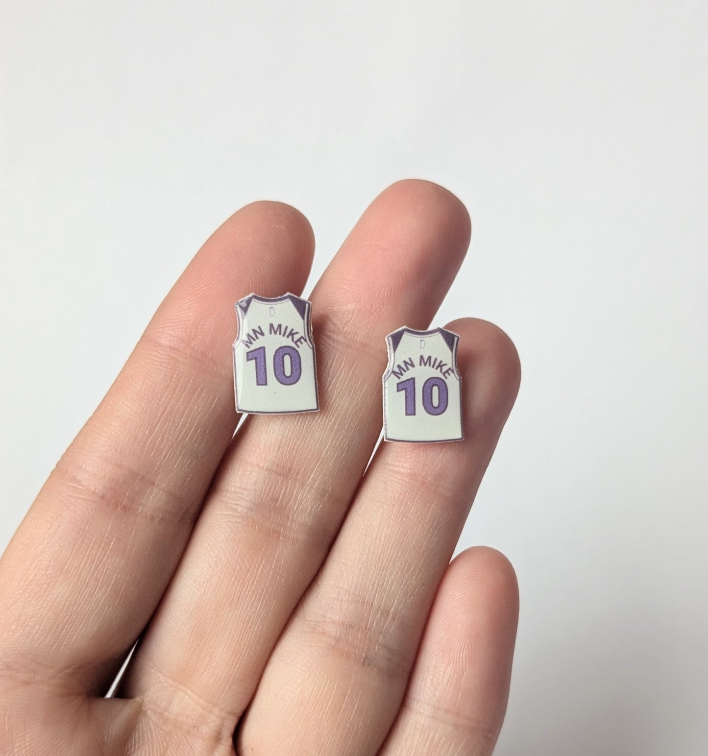 Two small earrings shaped like miniature jerseys with 'MN MIKE' and number '10' on a hand against a white background.