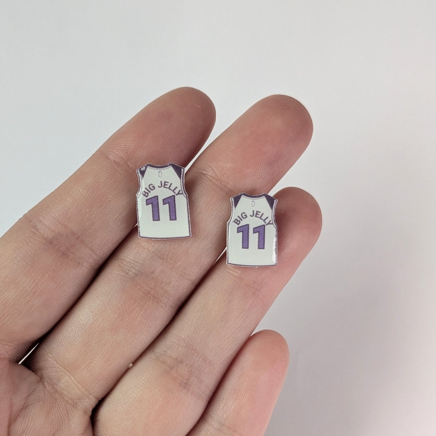 Two small earrings shaped like miniature sports jerseys held between fingers on a light background