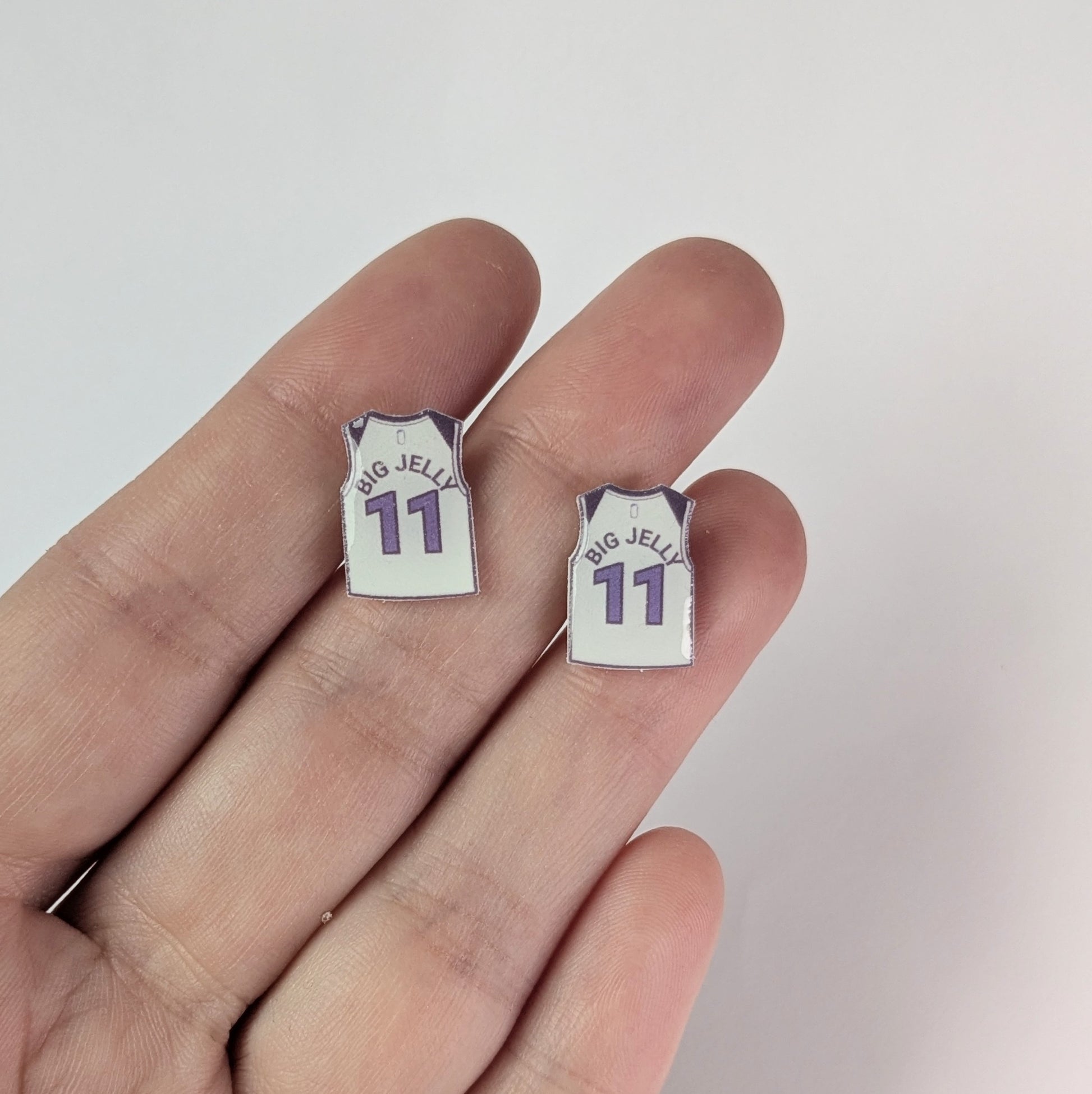 Two small earrings shaped like miniature sports jerseys held between fingers on a light background