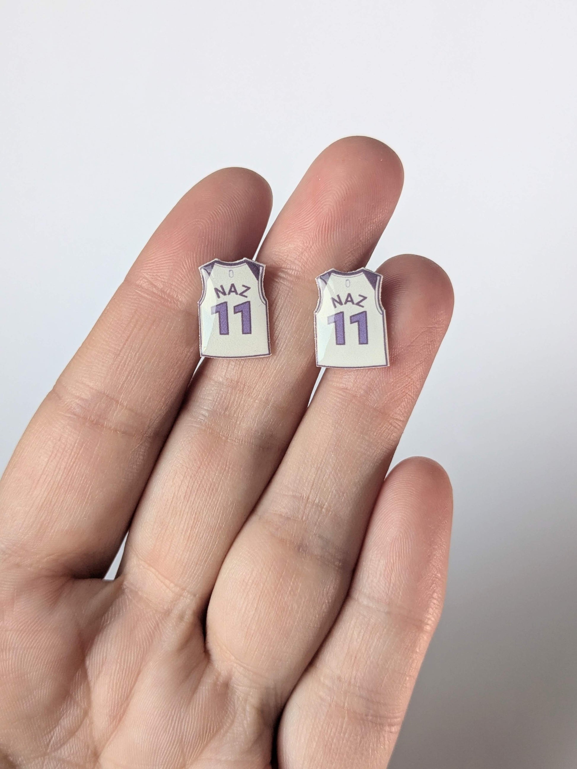 Two small earrings shaped like miniature sports jerseys with 'NAZ' and number '11' on a hand against a light background.