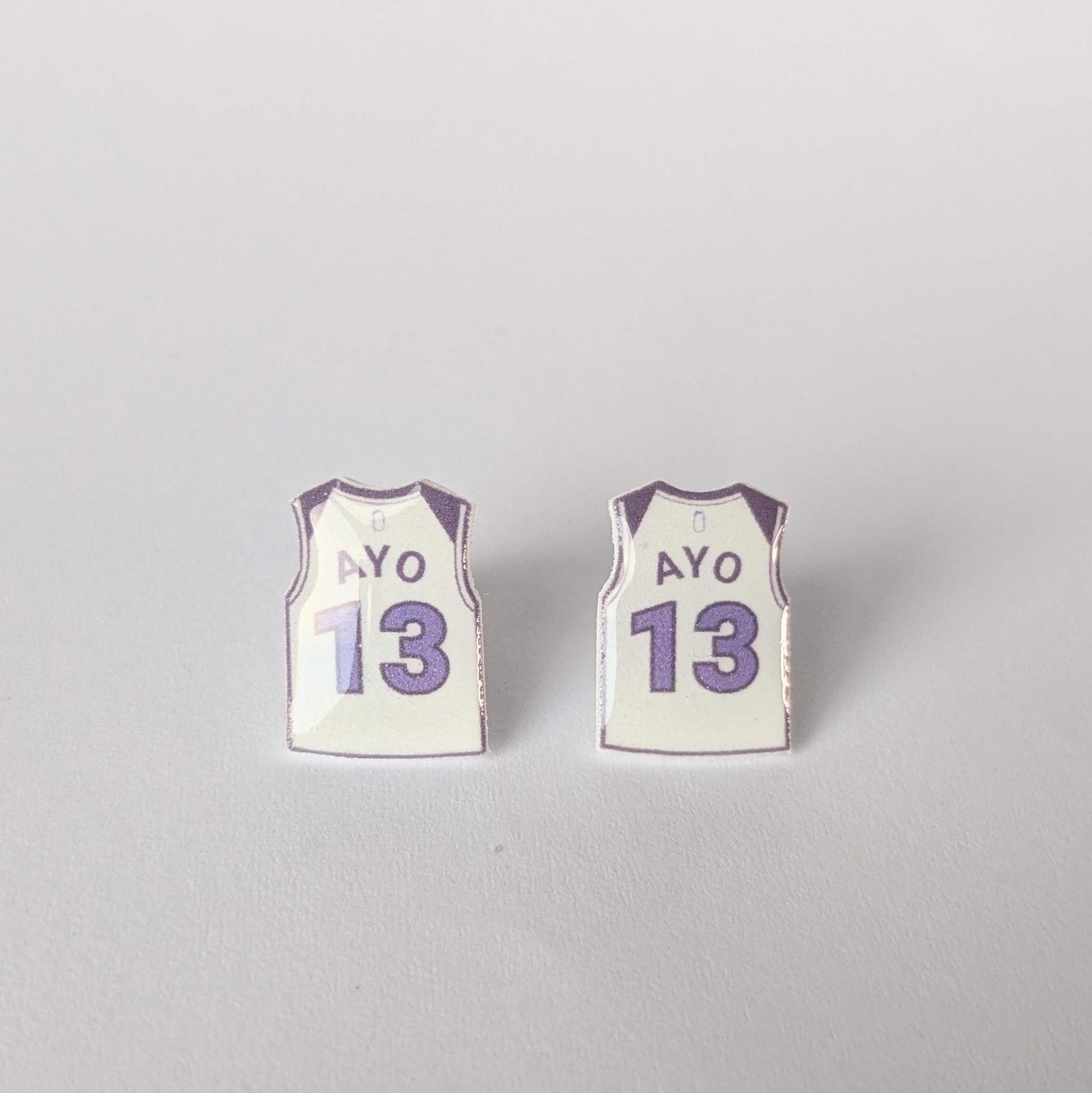 Two small earrings shaped like miniature basketball jerseys with 'AYO' and number '13' on a light gray background.