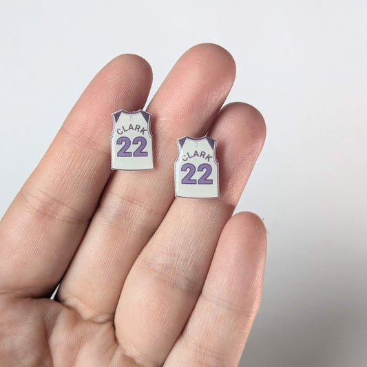 Two small earrings shaped like miniature sports jerseys with 'Clark' and number '22' on a hand against a light background.