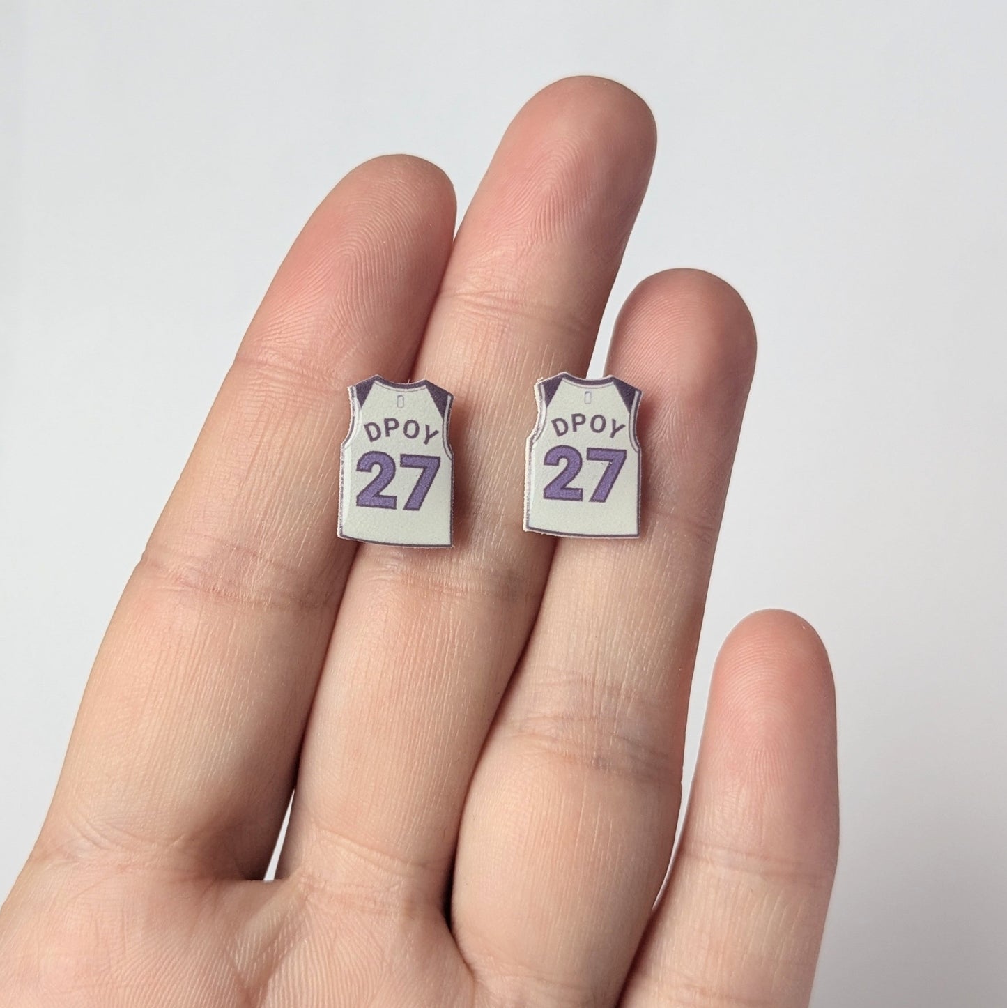 Two small earrings with 'DPOY 27' text held between fingers against a white background