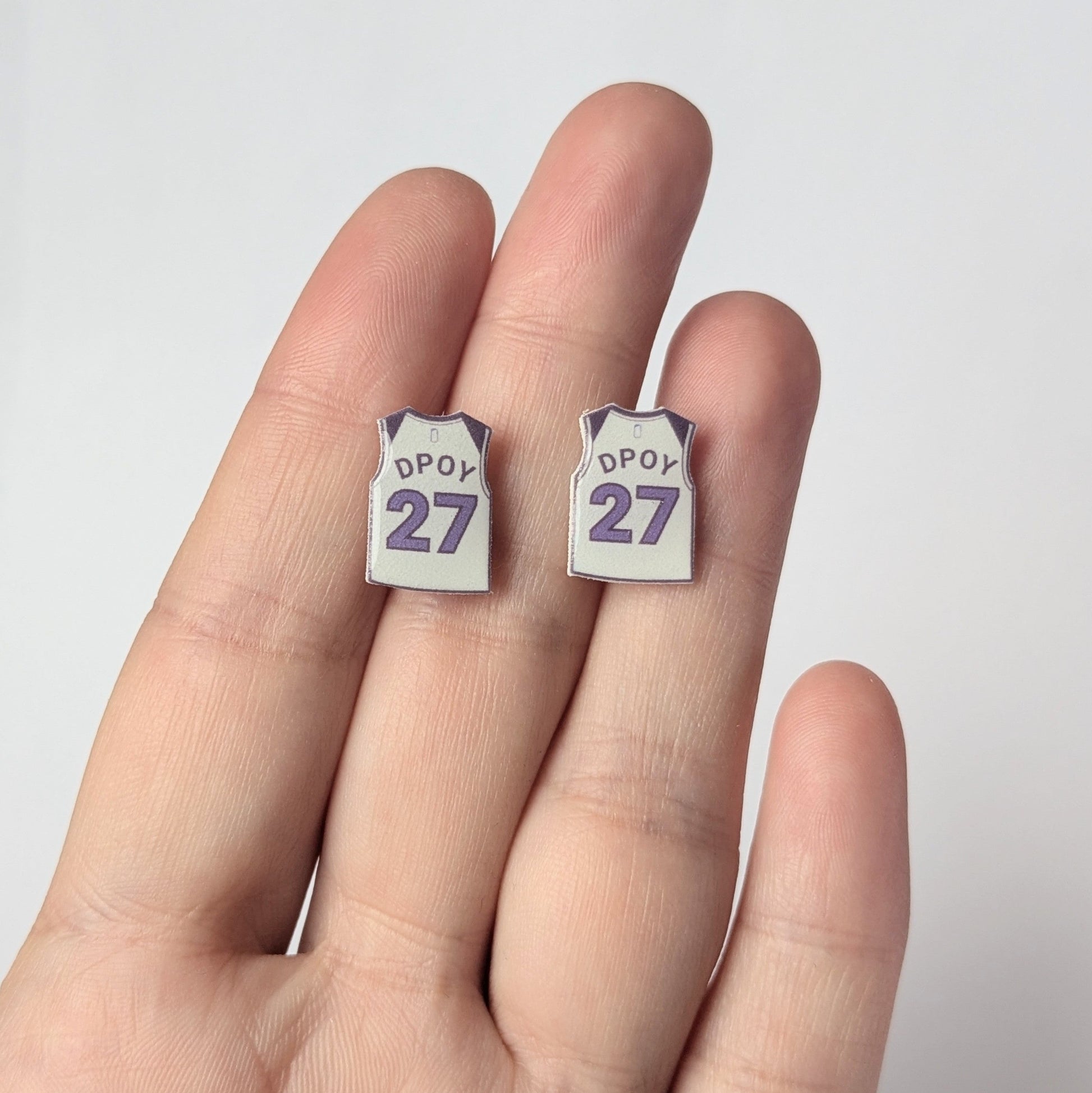Two small earrings with 'DPOY 27' text held between fingers against a white background