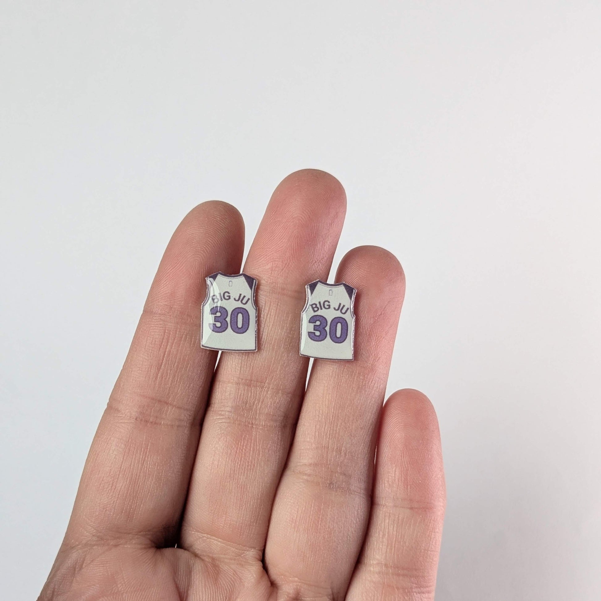 Earrings shaped like basketball jerseys with 'BIG JU 30' text held between fingers on a white background