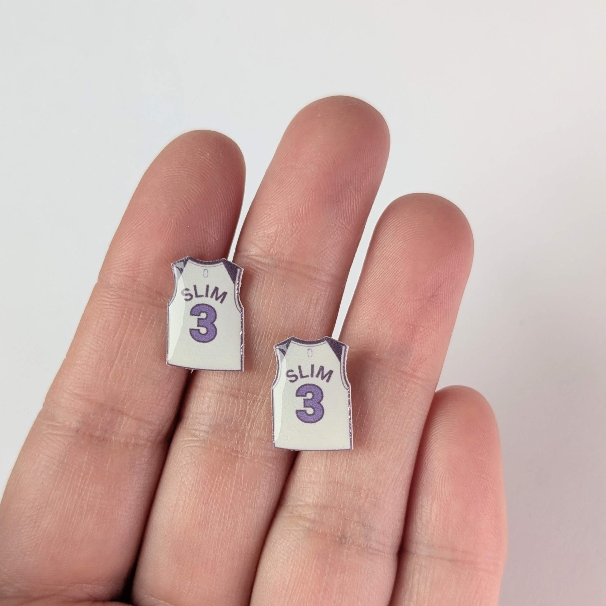 Two small earrings shaped like miniature sports jerseys on a person's finger.