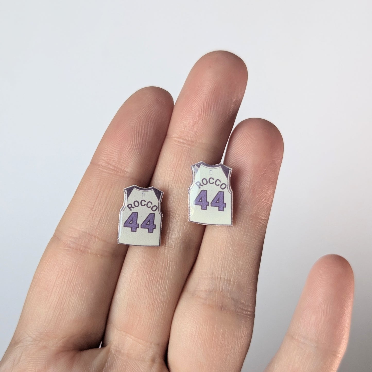 Small earrings shaped like basketball jerseys with 'Rocco 44' text on a hand against a light background