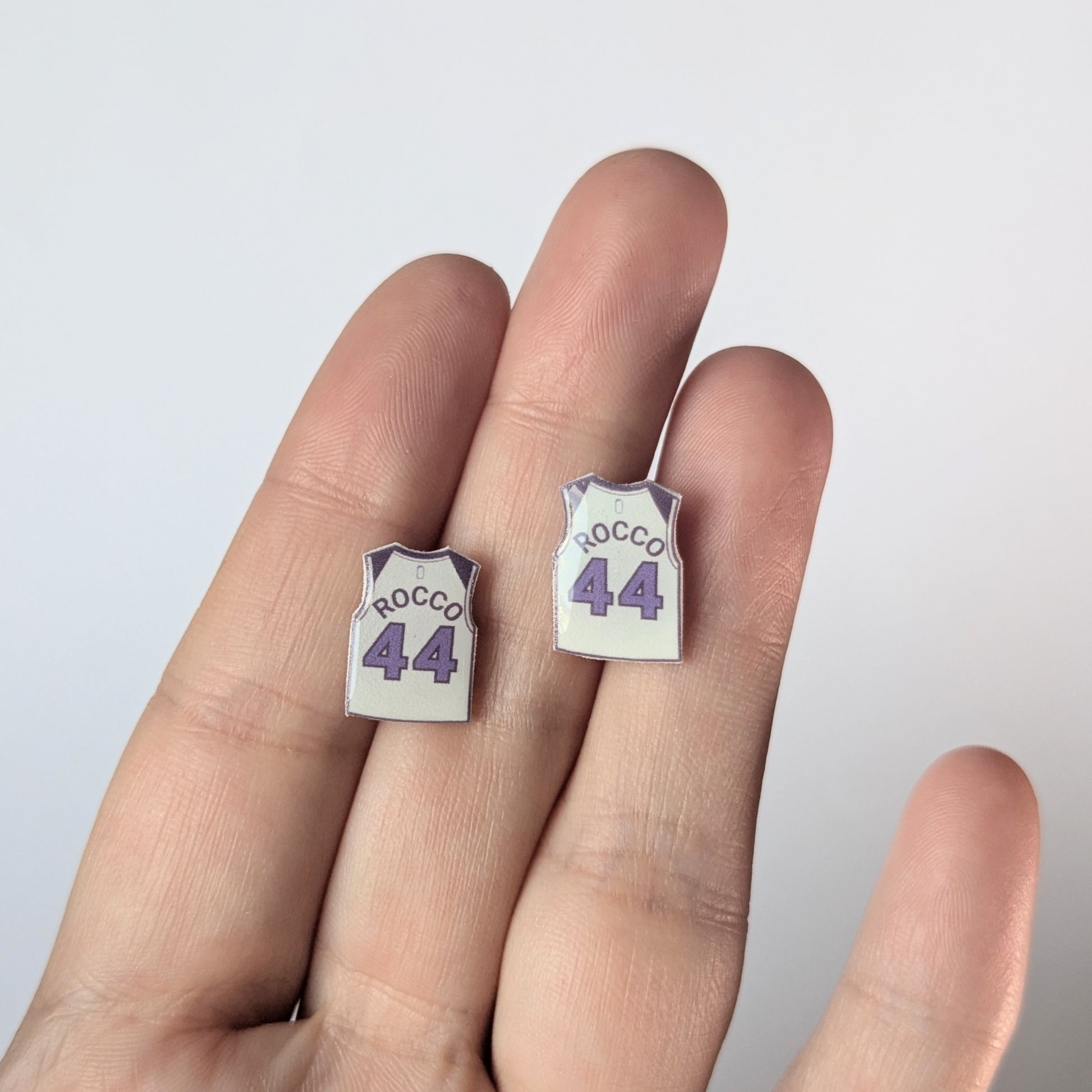 Small earrings shaped like basketball jerseys with 'Rocco 44' text on a hand against a light background