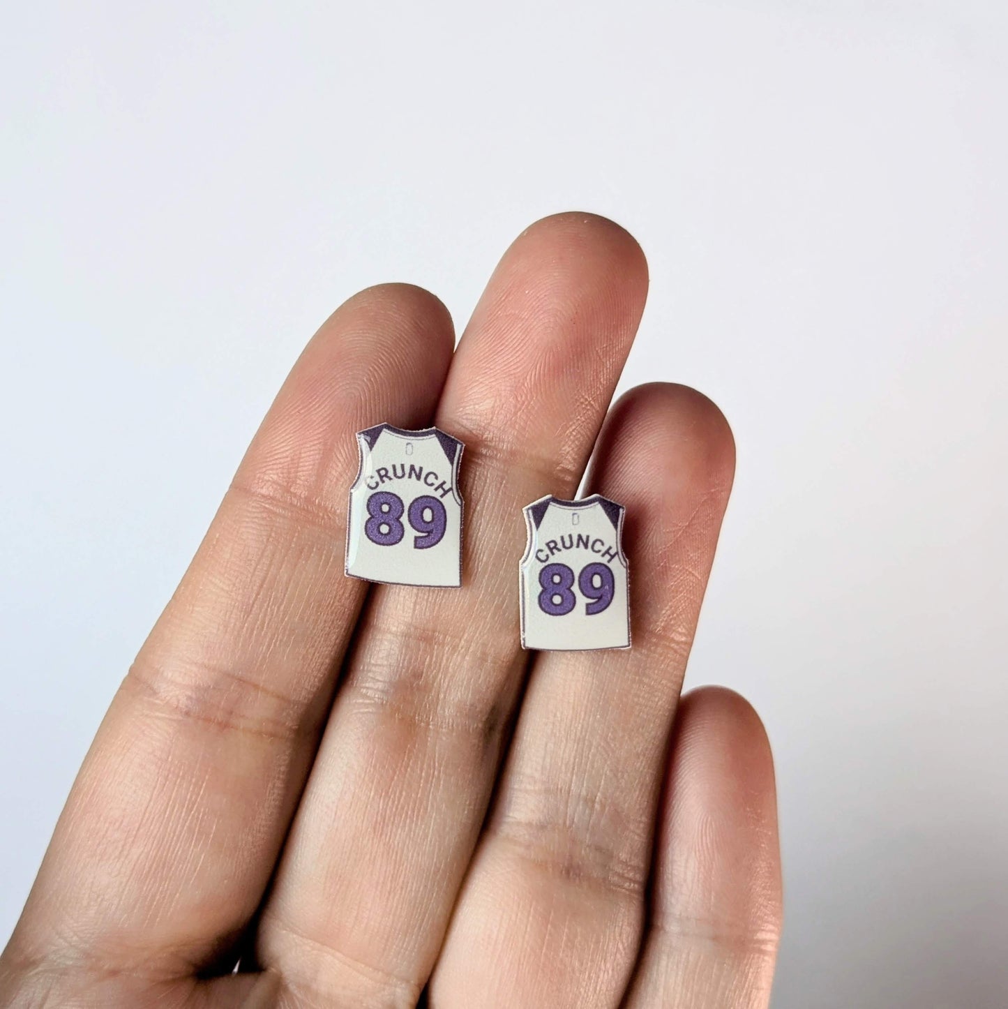 Two earrings shaped like miniature jerseys with 'CRUNCH 89' text on a hand against a white background.