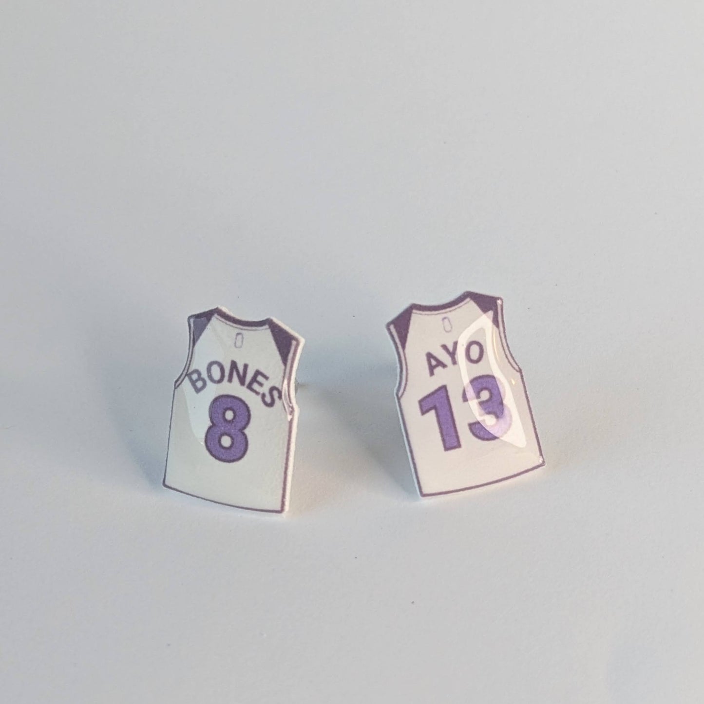 Two small earrings shaped like miniature sports jerseys on a light gray background