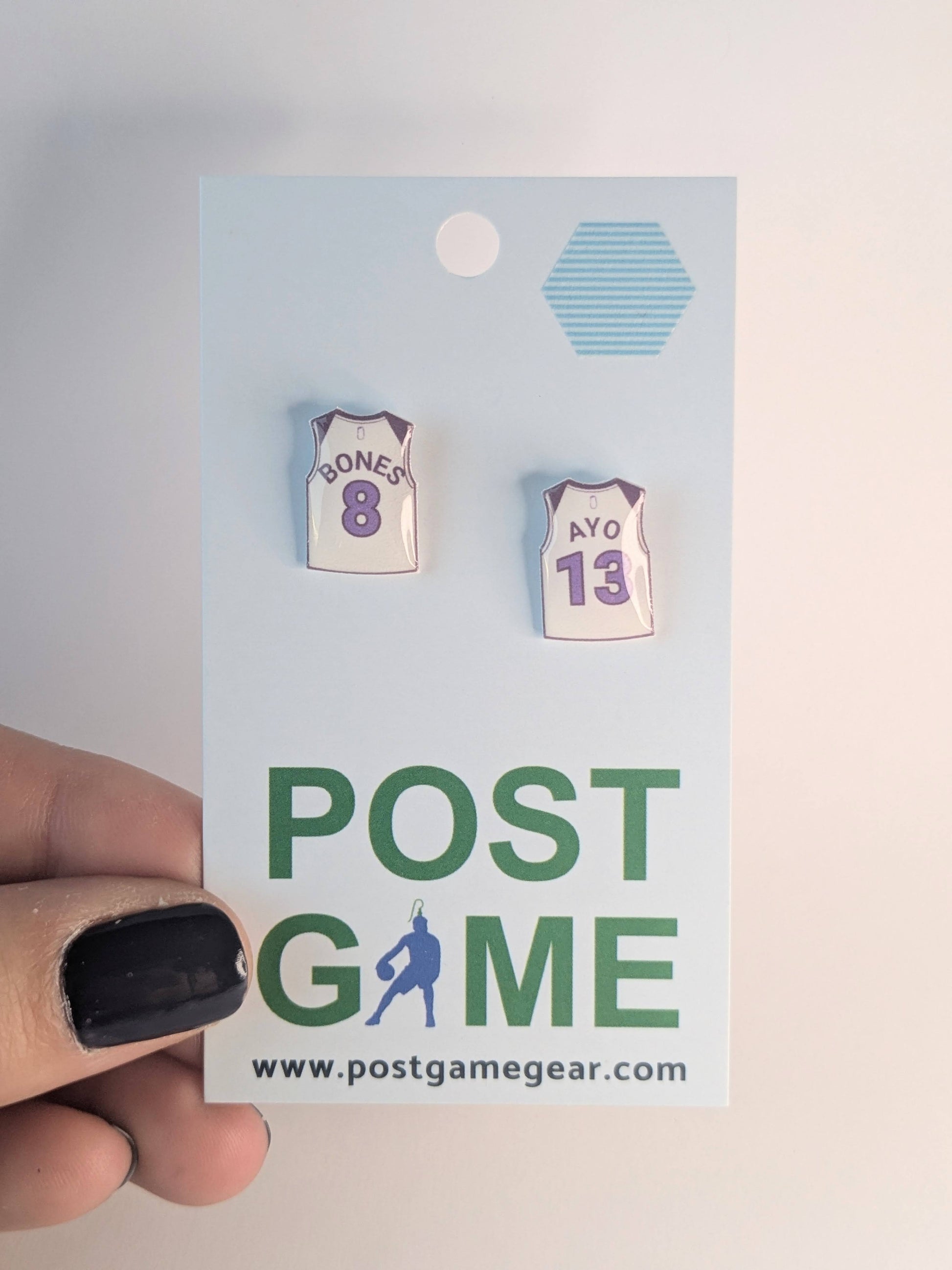 Two small earrings on a 'POST GAME' branded card held by a hand.