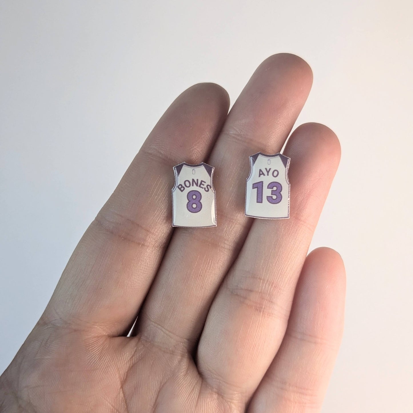 Two small earrings shaped like miniature basketball jerseys held between fingers on a light background